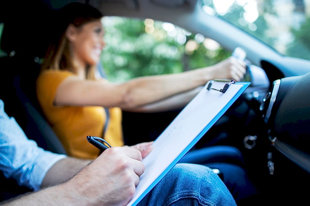 Close up view of driving instructor holding checklist while in background female student steering and driving car.