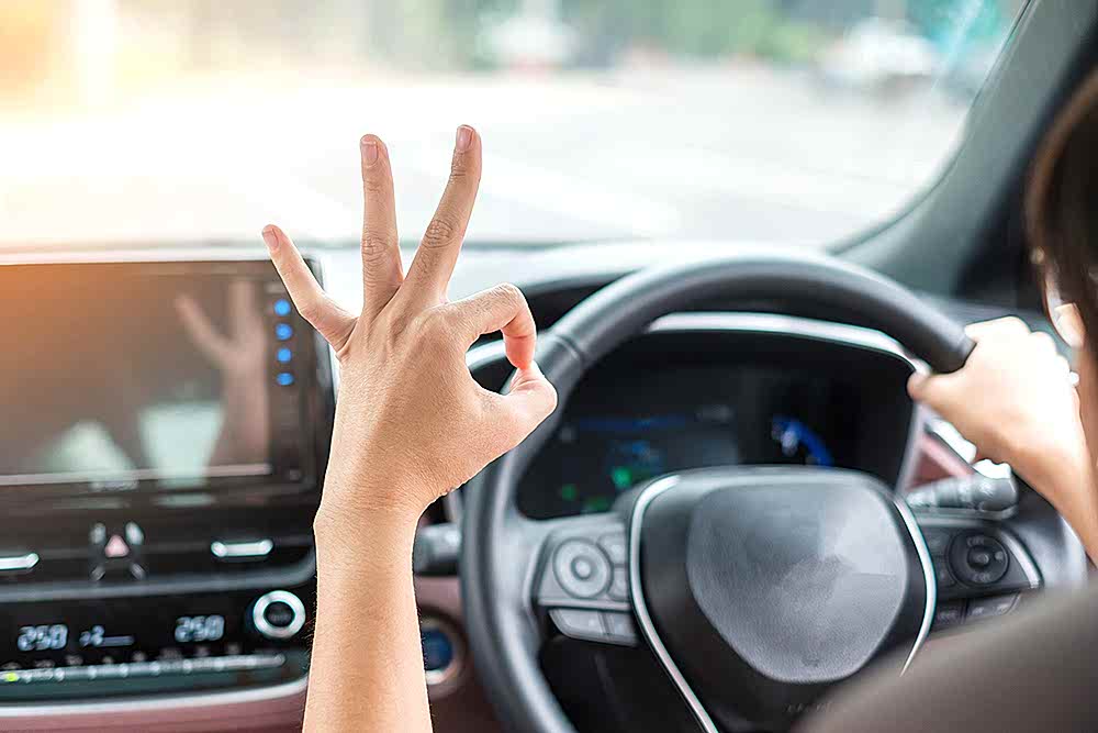 woman showing OK sign during driving a car on the road, hand controlling steering wheel in electric modern automobile. Journey, trip and safety Transportation concepts